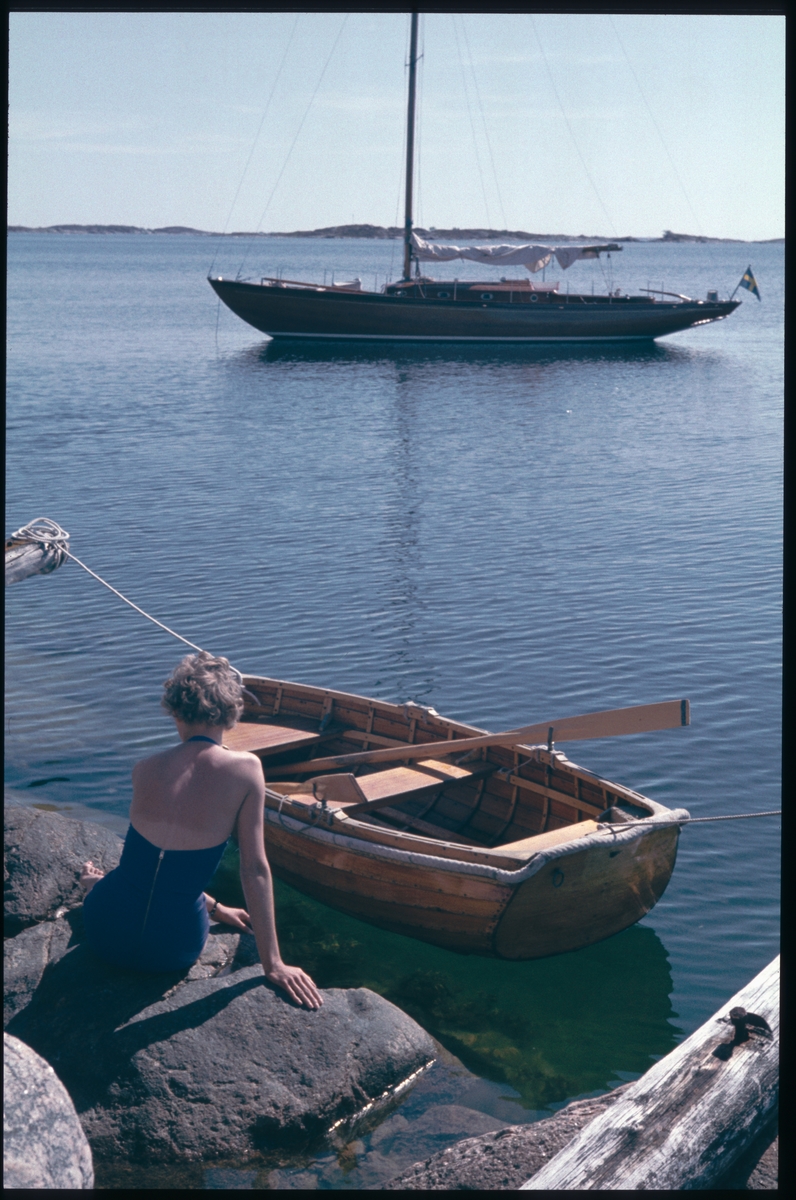 Kvinna med baddräkt på klipphäll i skärgården, segelbåten Elsi-Anne och en mindre roddbåt ligger förtöjda i närheten.