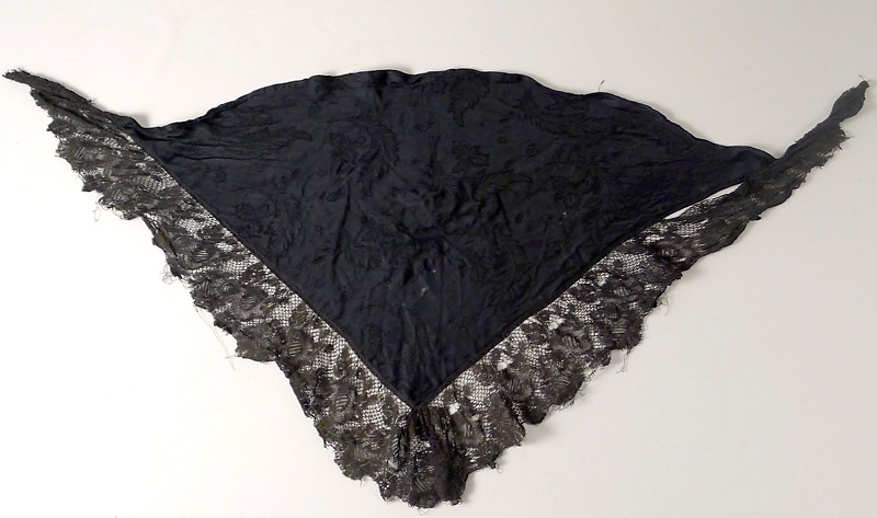 Trekantig sjal av konstsiden, med ca 8 cm bred maskintillverkad spets utefter kortsidorna. Sjalen är sannolikt en omgjord fyrkantig sjal. I mitten finns en laglapp av samma tyg.