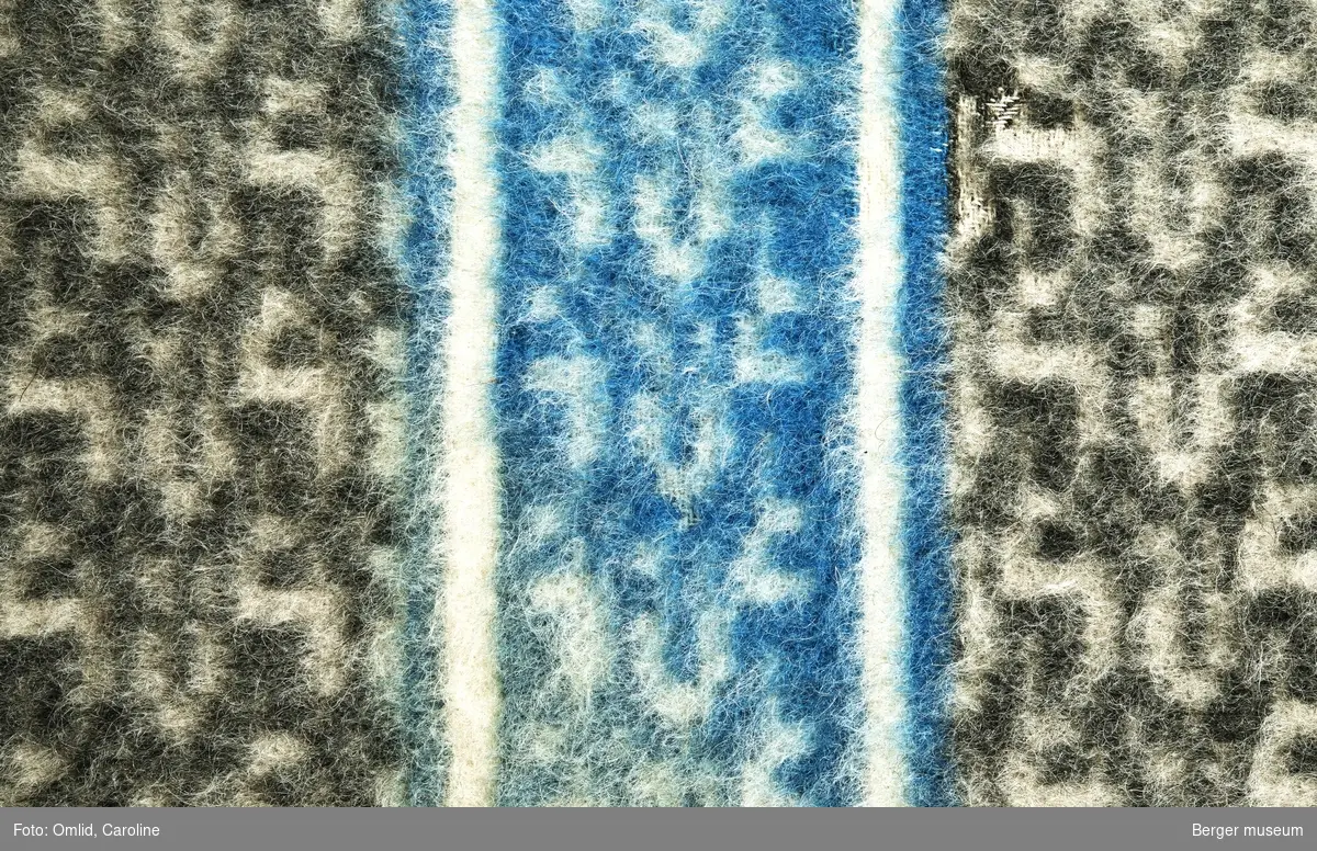 Teppe med geometrisk strukturmønster i mørk grått på lysere bakgrunn. Tverrgående blå bord med samme mønster.