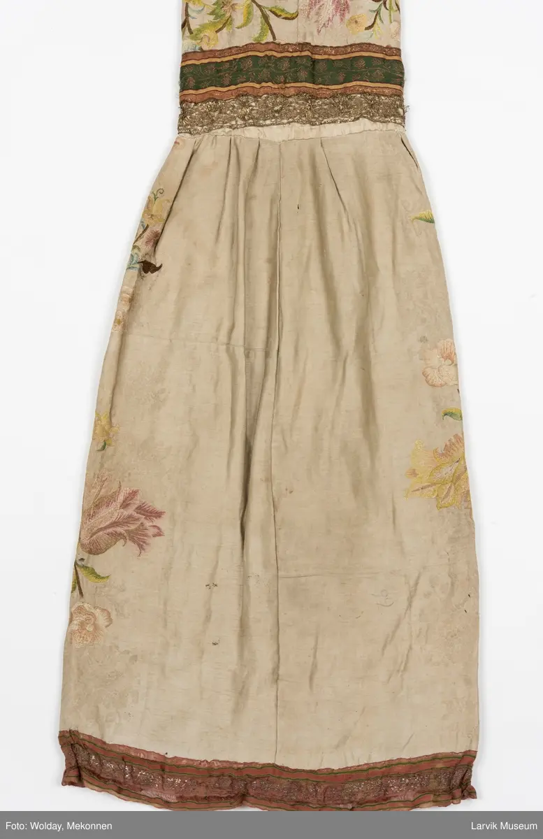 Form: livstykke som hette, åpen til livet, bred linning, rynket skjørt med blondekant nederst, søm bak på hetten i linning, søm foran og bak i skjørt, enkelte skjøtesømmer, linning brodert med gulltråd, skjørtet er foret med tynt stoff