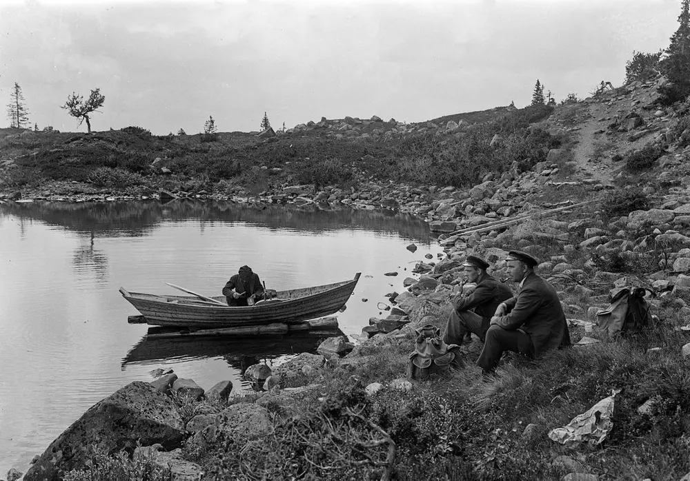 Lita bukt med en åfløy (robåt) som ligger ved land med én mann oppi, flere mennesker sitter på land, litt flatehogstaktiv landskap i bakgrunnen.