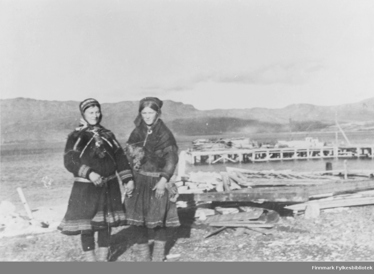 Reprobilde fra Nordreisa, Sørstraumen. Bilde tatt av et havneområde. To samiske kvinner i kofte står oppstilt i forkant av bildet. I bakgrunnen ser man et kaianlegg. Byggematerialer ligger rundt omkring i landskapet.