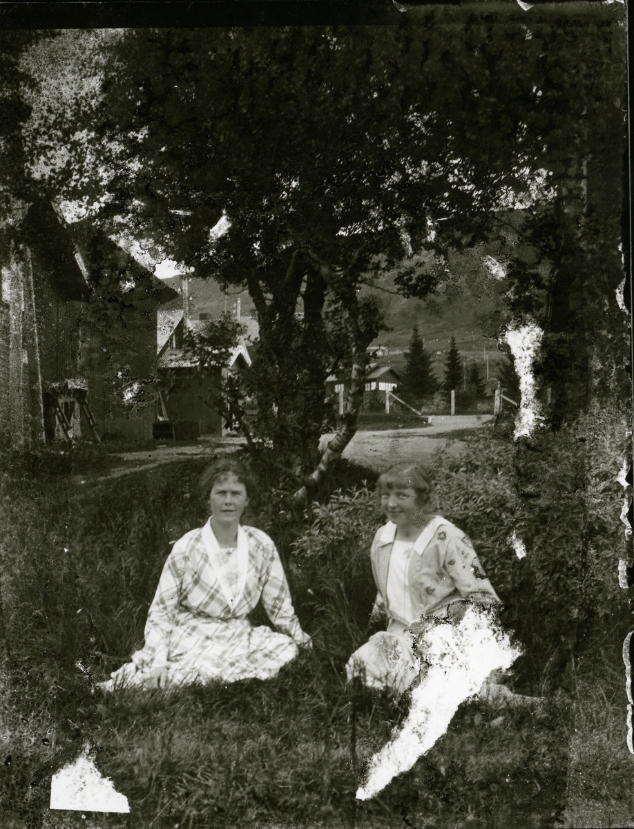 To kvinner sitter i graset foran et tre. Begge har kjoler.