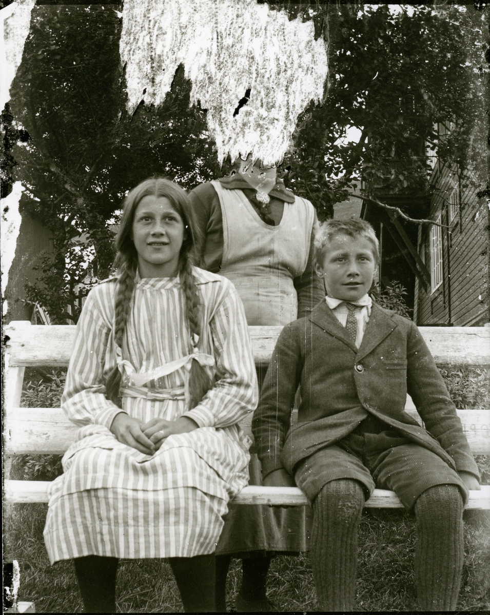 Jente og gutt sitter på en benk ute. Bak dem står en kvinne .