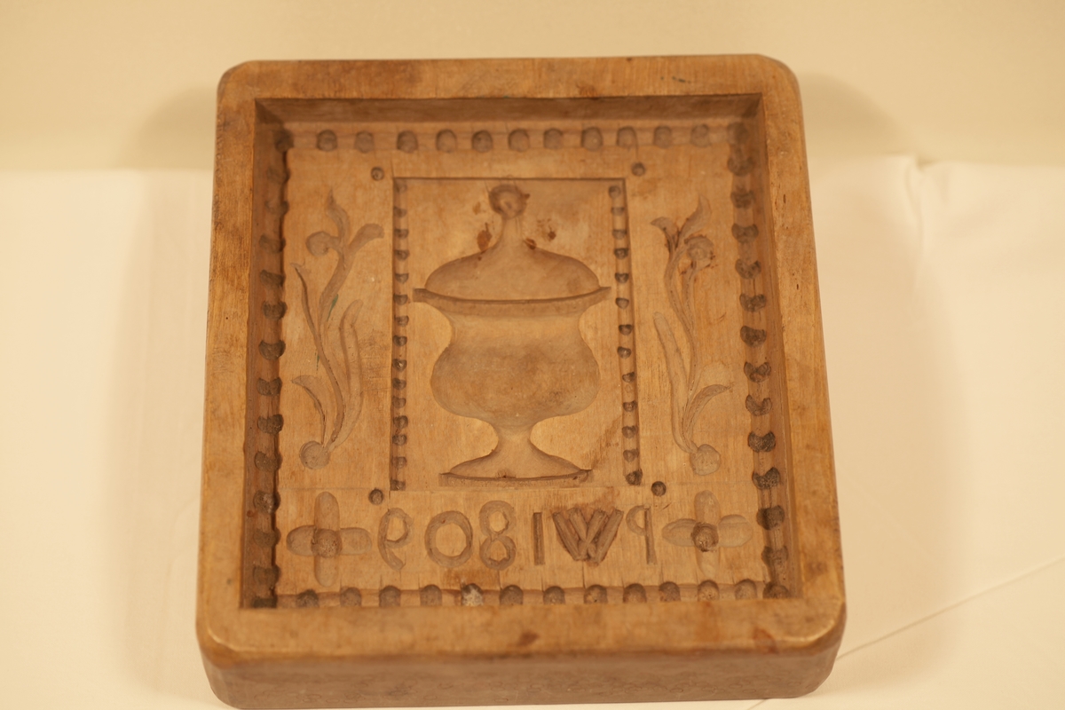 Form i tre. Utskjært motiv viser urne, blader og blomster.  Formens bakside er dekorert med hjortedyr og sidene med sirkler.