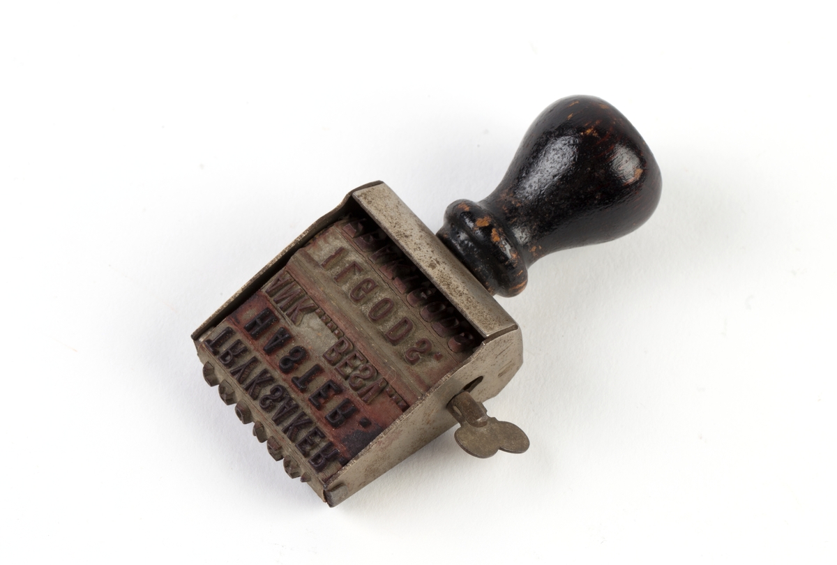 Stempel av stål med håndtak av sortmalt tre. Stempel av gummi som inneholder rader av tekst.
Hver enkelt tekstrad kan benyttes  ved  å dreie på en skrue på den ene siden av stempelet.