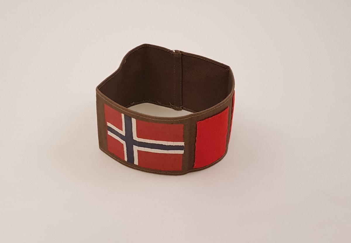 Brunt armbind påmalt et norsk flagg og påsydd en bred og en smal rød stolpe.