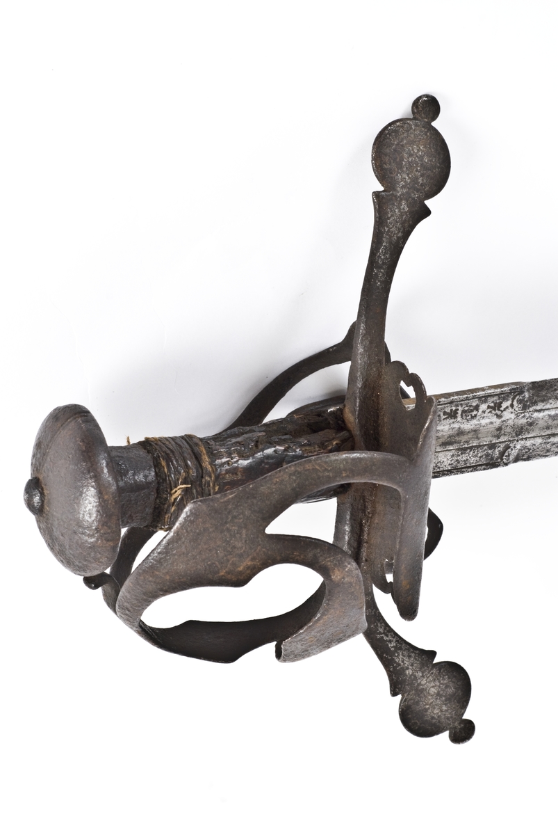 Rund soppformet knapp med nitteknapp  Tregrep med rester av hyssing bevikling. Flate svakt nedbøyde  parerstenger med som ender i to skiver. Punslet dekor på parerstengene.
Asymmetrisk kurvfeste med nyreformet parerplate på utsiden og klingebrekker på innsiden. Sidebøyler og håmdbøyle er formet av flate bånd.
Tveegget rett klinge med  6 kantet tverrsnitt  og en  midtstillt kort blodrand. Klingen er merket med diverse smedstempler og bokstaver.