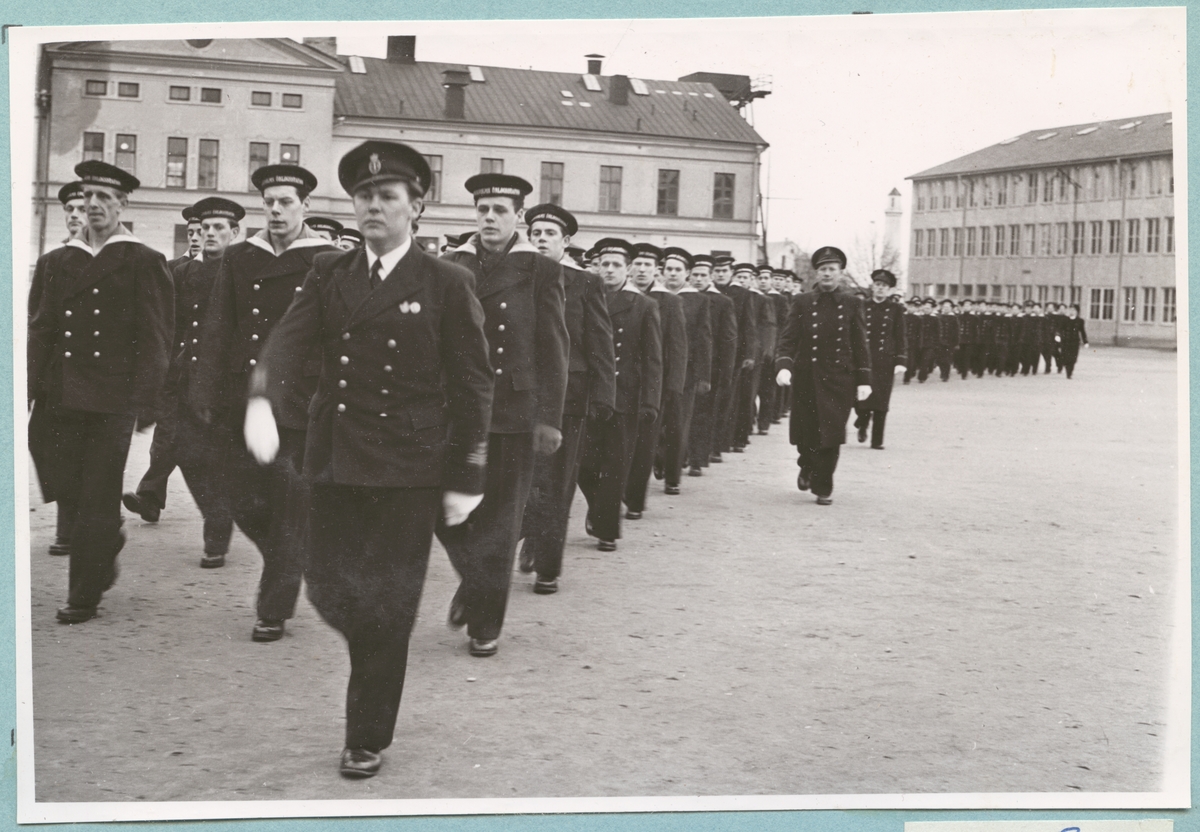 Uniformsklädda flottister marscherar över bataljon Sparres kaserngård. I bakgrunden syns matinrättningen med putsad fasad och plåttak.