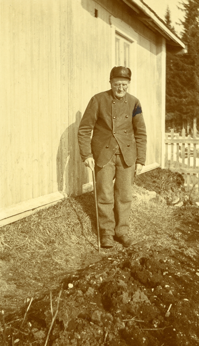 Gammel mann med briller og stav. Stående ved husvegg. Paul Gregersen, søndre Sørlie, Langtjernåsen (28/12 1847 - 15/11 1928) (78 år gammel)