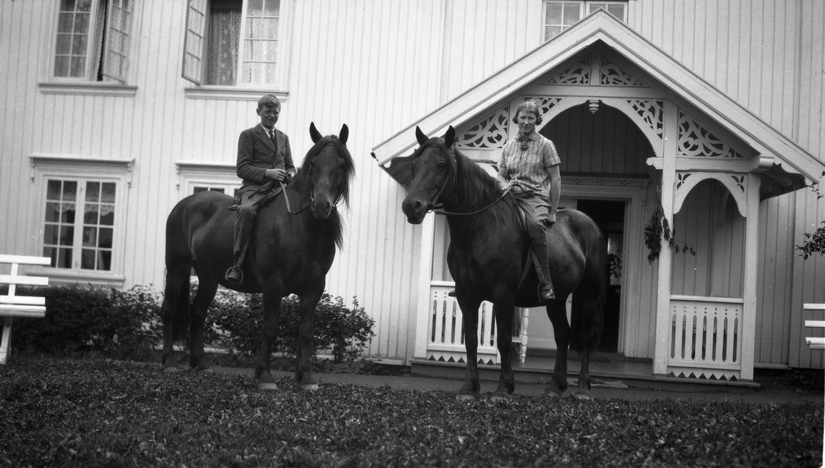 To uidentifiserte ryttere, mann og kvinne, foran inngangspartiet til et våningshus. Stedet er ikke sikkert identifisert, men kan muligens være gården Hval i Østre Toten. To nær identiske bilder.