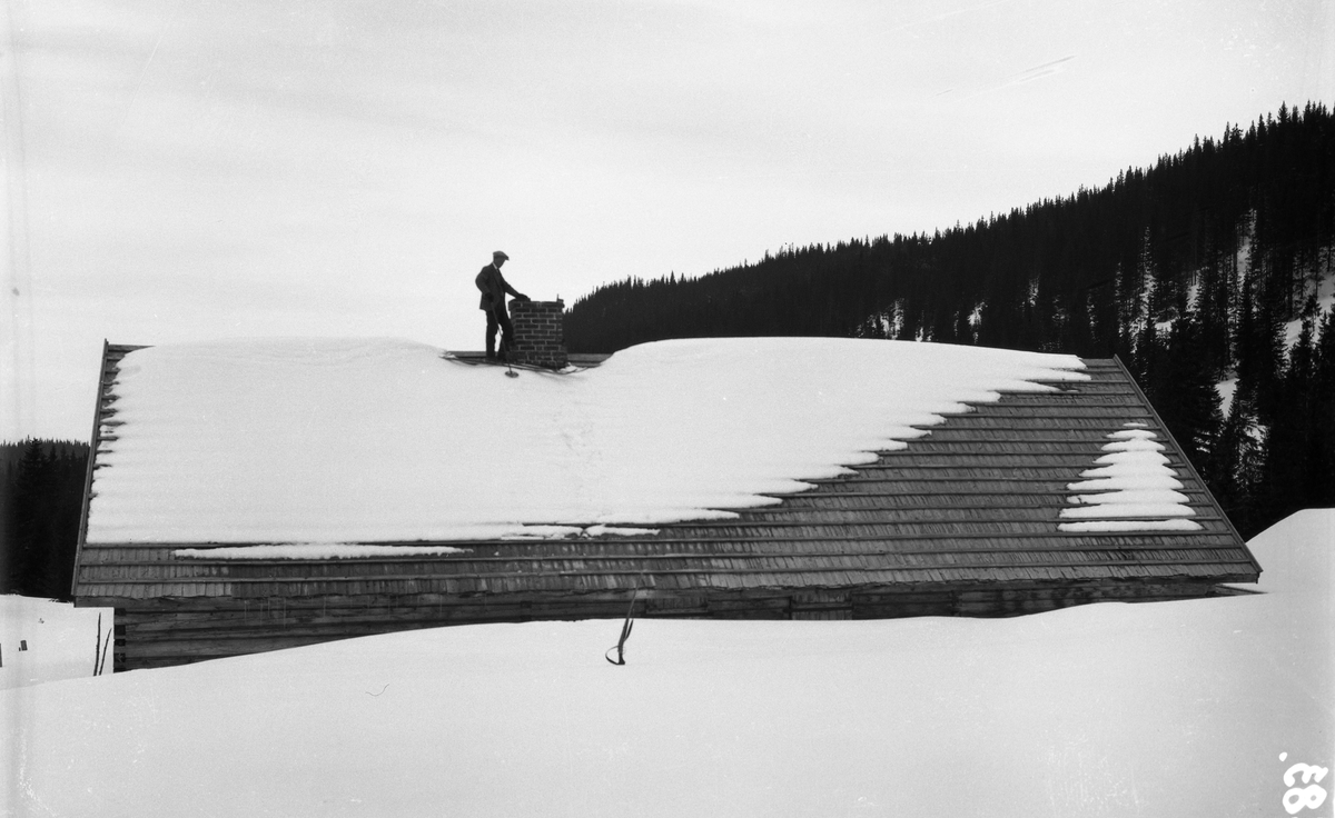 Snøvinter. En skiløper står på taket av noe som trolig er et seterstørhus. Hverken person eller sted er identifisert.