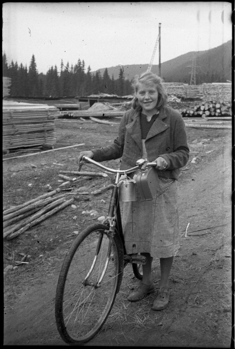 Uidentifisert ung kvinne med sykkel. I bakgrunnen synes å være et sagbruk. Stedet er ikke identifisert.