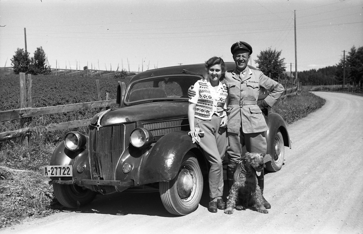 Major Bjart Ording og en uidentifisert kvinne ved en bil. Bilen er iflg. informant en Ford V8 1936 modell. Den mangler støtfanger foran, og har også andre skader i fronten.