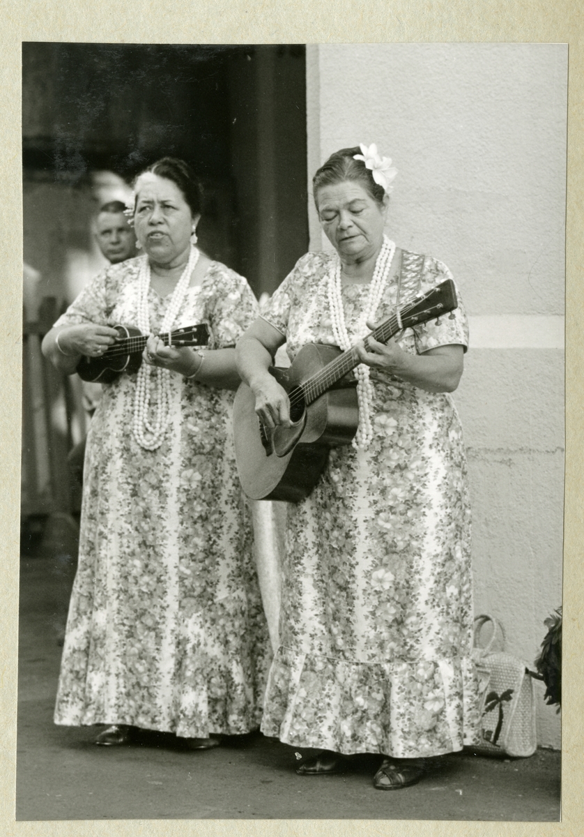 Bilden föreställer två kvinnor som spelar gitarr och ukulele. Båda är klädda i klänning och är utsmyckade med halsband och blommor. Bilden är tagen under minfartyget Älvsnabbens långresa 1966-1967.