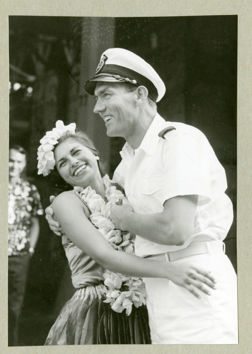 Bilden föreställer en besättningsmedlem i vit uniform som omfamnar en kvinna utsmyckad med blommor i håret och runt halsen. Bilden är tagen i samband med minfartyget Älvsnabbens långresa 1966-1967.