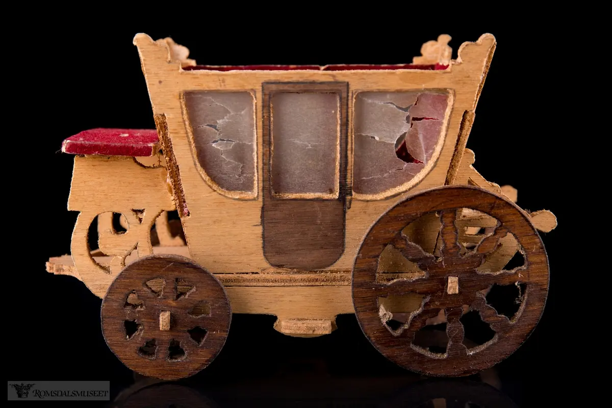 En liten leke- hestevogn i kryssfiner. To store og to små hjul, kledd med rød filt og vokspapir er brukt som vinduer.