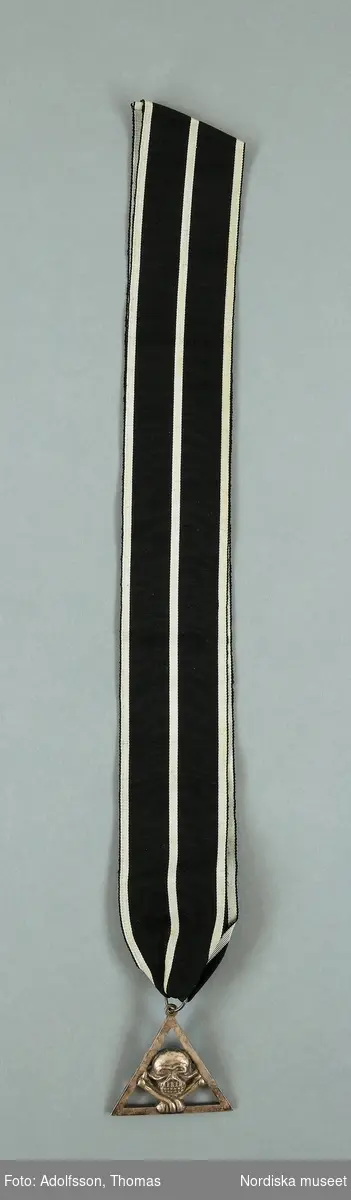 Huvudliggaren 1896:
"c. L. 0,051 B. 0,058, Triangel af hvit metall inuti triangeln en dödskalle och två korslagda benkotor.
I svart sidenmoaréband, med hvita ränder. Bars om halsen."