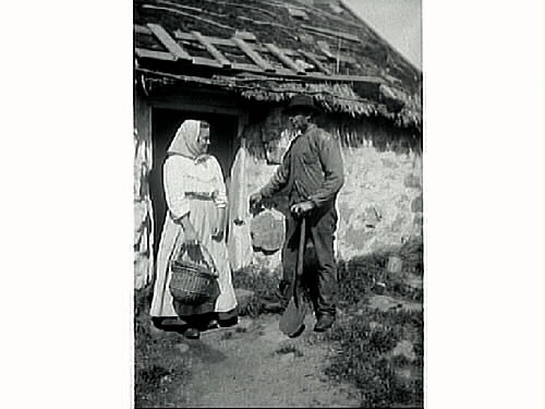En arbetsklädd man med spade och en ung kvinna med korgar framför dörren till en stenstuga med halmtak.