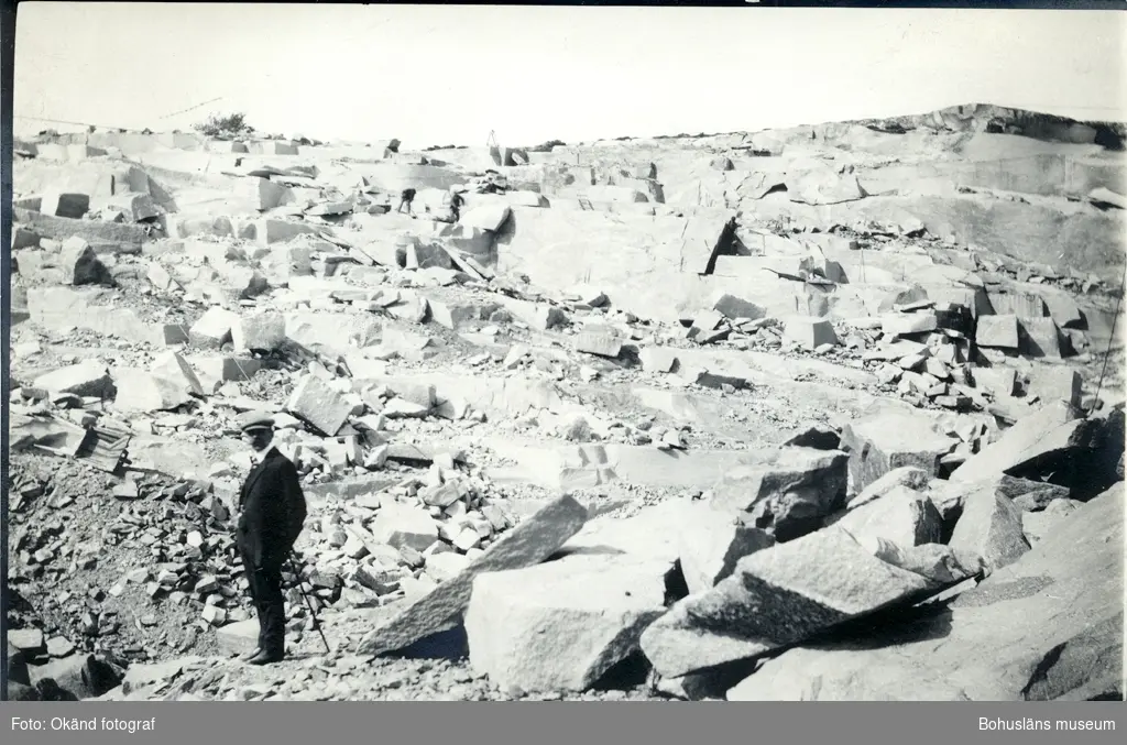 En man i kostym och med spatserkäpp står i stenbrotten på Rixö, han tittar in i kameran. Det är soligt och i bakgrunden arbetar två personer uppe på stenblocken