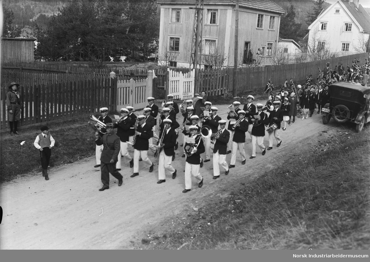 17. mai feiring 1933. Korps marsjerer i gate, barn med flagg i hånda gående bak. Bil parkert lang gaten. Bolighus med mennesker stående på veranda. Tilskuere langs gaten.