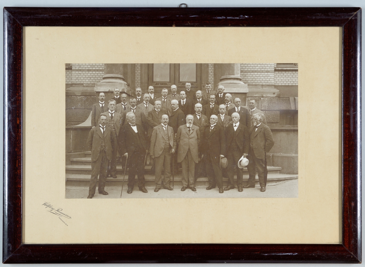 Svart-hvitt fotografi av deltagerne på Apotekermøtet i Trondhjem i 1918. Det er i alt 30 menn fordelt på 3-4 rekker som er avbildet. De står foran en murbygning i Trondheim.