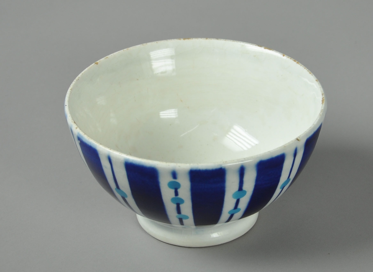 Hvit skål av glassert keramikk, med påmalt dekor. Motiv på malt dekor er striper og sirkler.