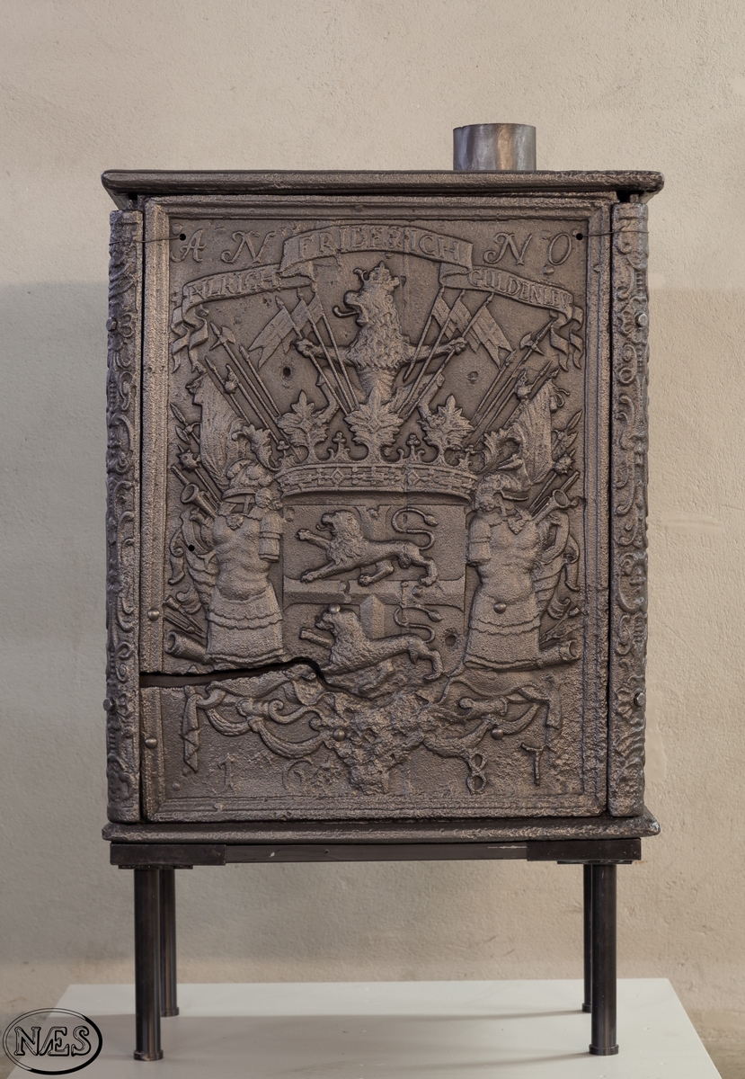 Kasseovn med ovnsdør på kortsiden og røykuttak i topplaten. Ovnen har løse skinner med bruskornamenter. På langsidene er Gyldenløves våpen omgitt av krigerske emblemer med innskrift i knekket bånd over. Øverst står det innskrevet ANNO og nederst på platen 1687. På kortsiden er det øverst krigerske emblemer og nederst den norske løve omkranset av brusk-kartusj.