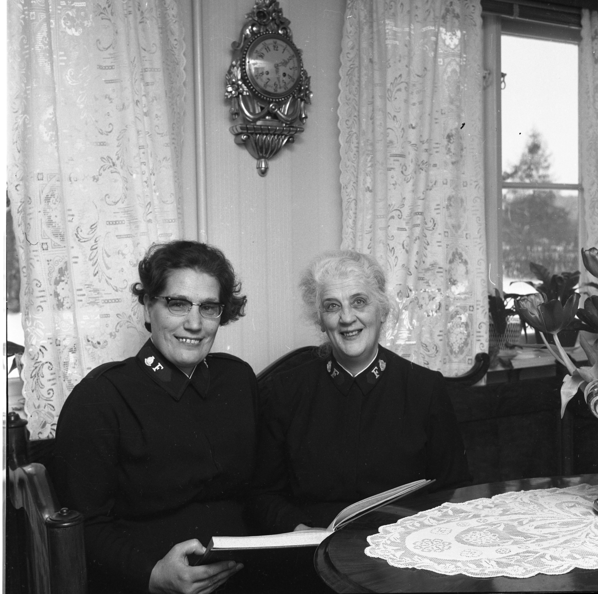 "Smålandsgården, vaktombyte". Två okända kvinnor i frälsningsarméns uniform sitter i en soffa med en uppslagen bok i händerna.