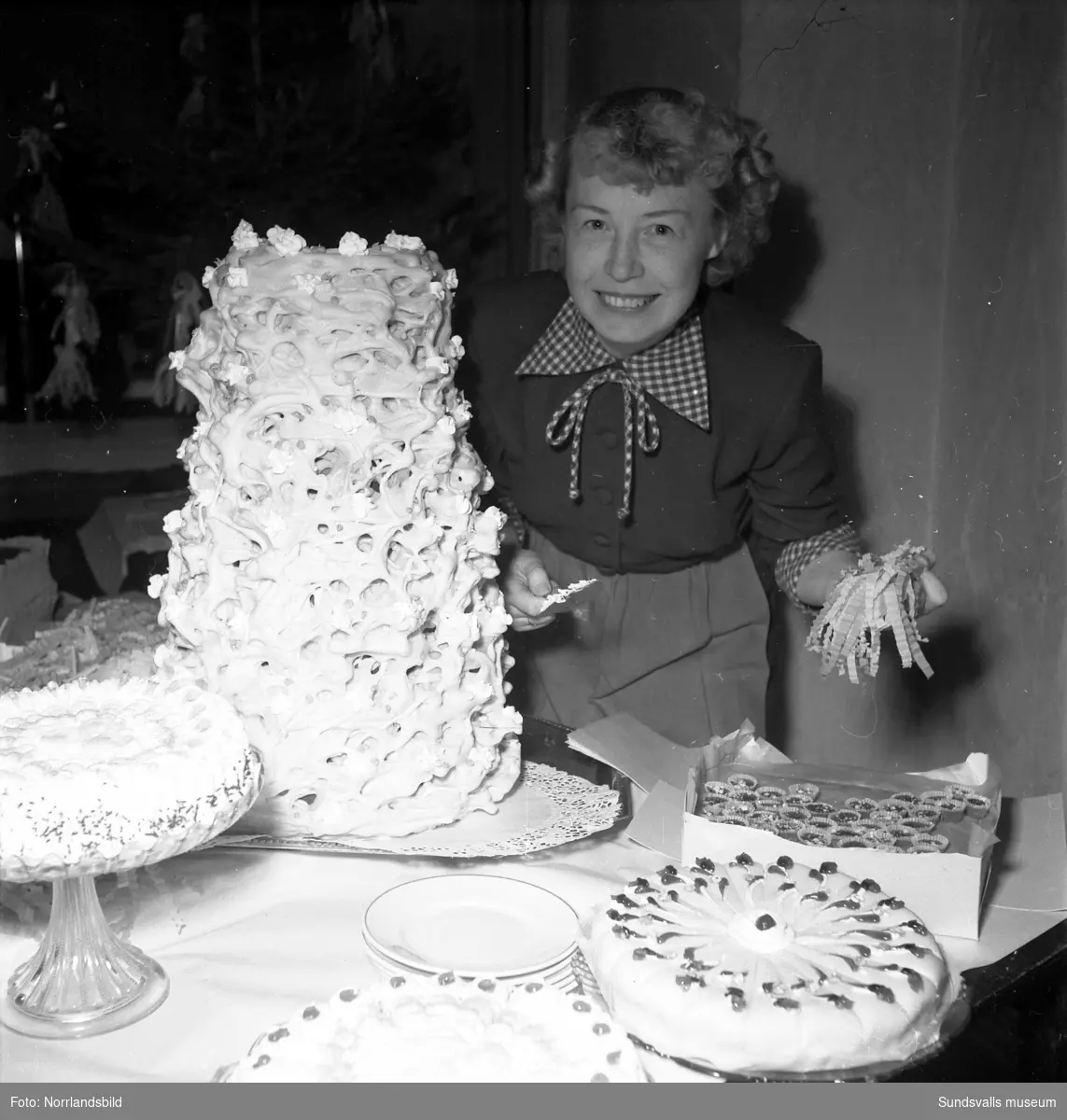 Hörselfrämjandets mässa på Församlingshuset. Kvinnor vid ett bord fullt av tårtor och andra godsaker, bland annat en imponerande spettekaka. En flicka och ett dockskåp.