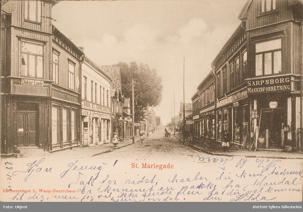 Postkort, lystrykk av St.Mariegate i Sarpsborg , datert 1903. 
Diverse reklameskilt: 
Doktor Lande, Sarpsborg Maskinforretning, Chr. E. Greaaker, cycle og uhrmagerforretning,