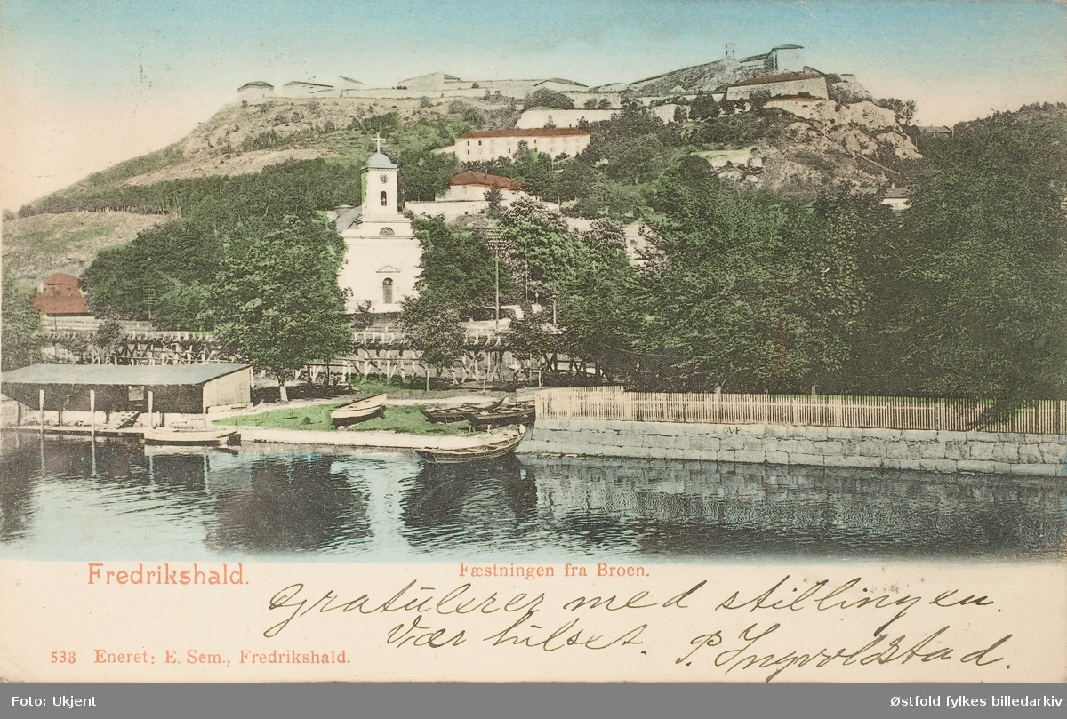 Postkort, kolorert lystrykk av parti fra hlden; Tista i forgrunnen Fredriksten festning og Immanuelskirken. Poststemplet 1904.