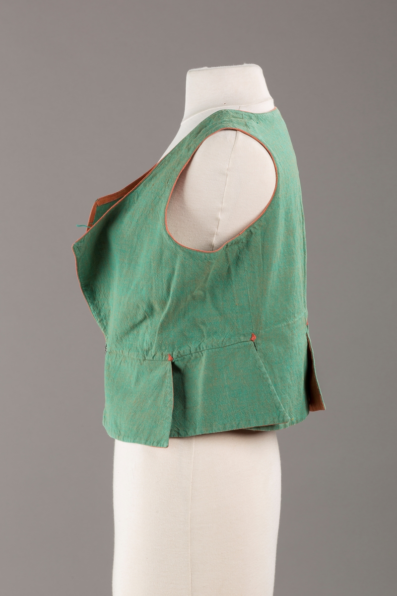 Grønt liv med rustrød kant og fór, seks skjøter. Håndsydd forsterkning i kontrastfarge øverst i skjøtesplittene. Lukkes med tinnhekte.