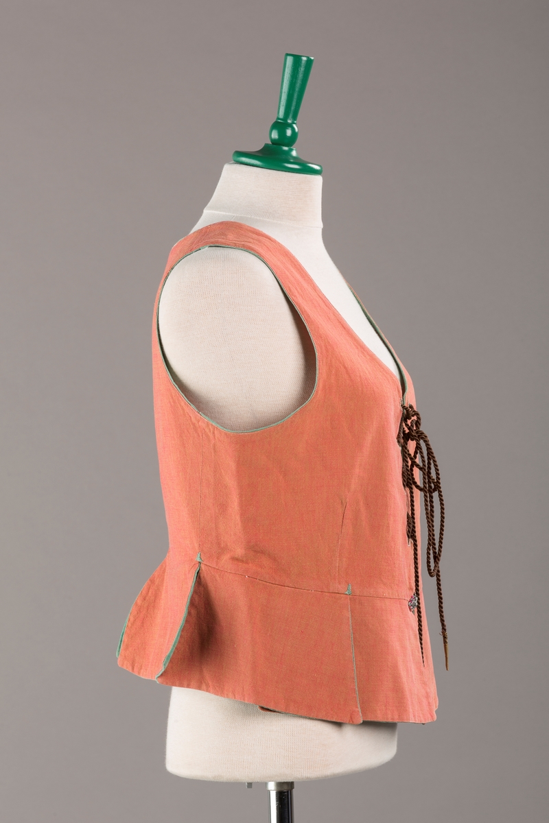 Rustrødt liv med grønn kant og fór, seks skjøter. Håndsydd forsterkning i kontrastfarge øverst i skjøtesplittene. Lukkes med tinnhekte og silkesnor.