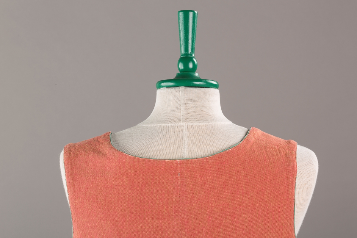 Rustrødt liv med grønn kant og fór, seks skjøter. Håndsydd forsterkning i kontrastfarge øverst i skjøtesplittene. Lukkes med tinnhekte og silkesnor.