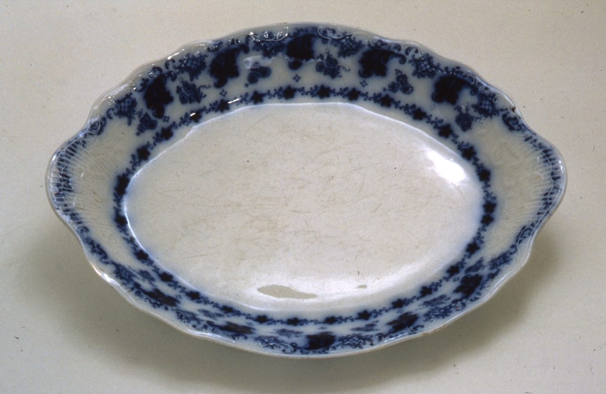 Ovalt fat i porselen med utoverbrettet tungekant. Fatet har blå dekor i form av blomster og bladverk. Dekor i godset.