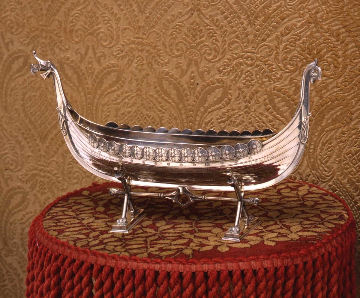 Støpt vikingeskip i sølv med dragehoder og skjold. Understellet har kryssende ben som ender i løveføtter, tversprosse og to profilerte fotstykker