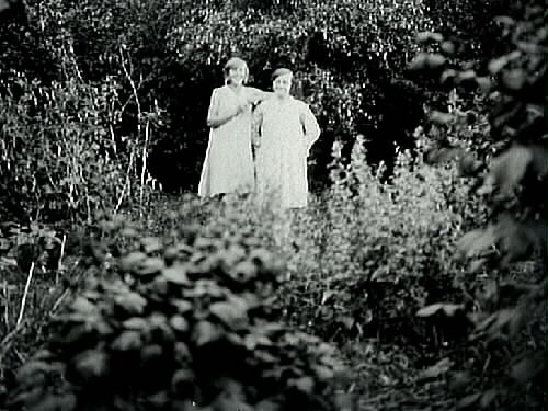 Två ledigt vitklädda kvinnor står i en frodig grönska.