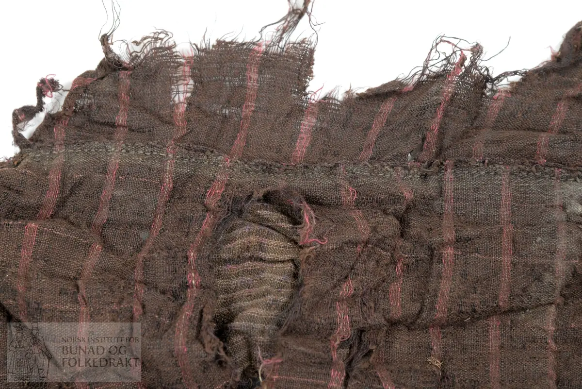 Svært utslitt del av ein stakk med linning, nederkant og ei metallhekte i eine enden av linning. Nedanfor denne delen av linning er det sauma fald som truleg er splitten midt framme på stakken. Av tre ulike verkensstoff, lerretsbinding med bomull i renning, ull i innslag. Stripete stoff i brun og lilla, svært utydeleg lillafargar. Fleire små hol etter mus, har vore nytta som stappefille.