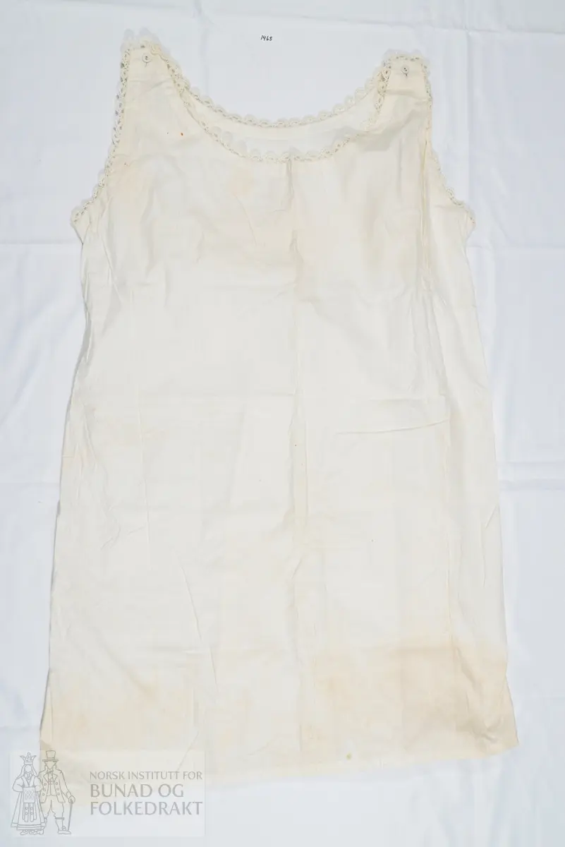 Nattkjole i bomullslerret med smal heklet kant rundt hals og ermringing. Sydd av et for- og bakstykke, med knepping med knapphull og knapp på skuldrene. Sydd sammen med indresøm på maskin. Svært små sting. Den påsydde blonden er heklet av mercerisert bomullsgarn. Dobbel oppbrett nederst på skjørtekanten.