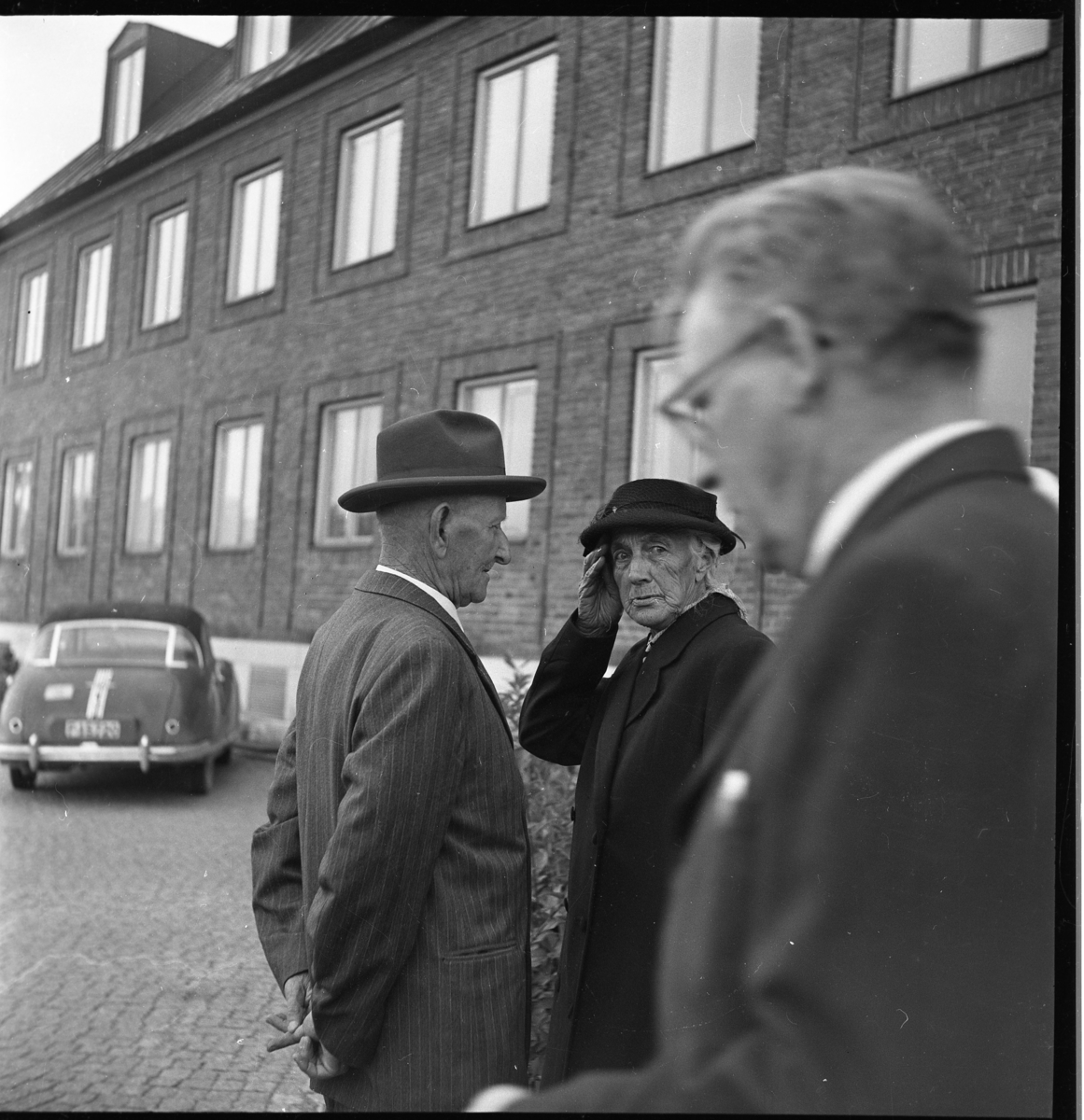 "Kalle Vacker" Karlsson i randig kostym och hatt intill äldre kvinna i kappa, hatt och med handväska. En okänd man kommer in i bild från höger.