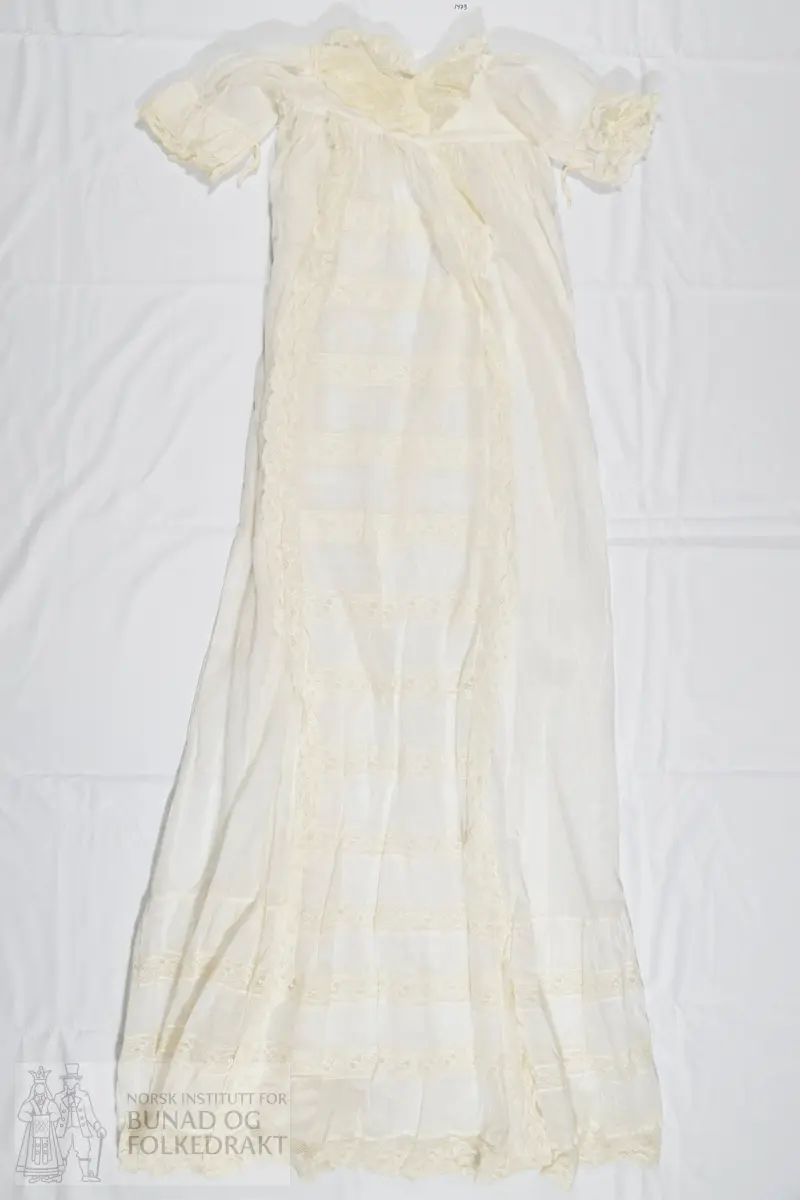 Lang dåpskjole i hvit bomullslerret med påsydde kjøpeblonder. Livstykket er sydd av to lag med tynn bomullslerret. Sydd til skjørtet med en bred linning. Ermer sydd rett i, med rynker langs hele ermeåpningen. 

Åpning midt bak med splitt et stykke ned på skjørtet, knyting øverst med bånd. Forstykke og de to bakstykkene er sydd sammen på skuldrene. Halslinning er brettet ned dobbelt på baksiden, og er sydd ned på maskin. Bred blonde i halsringingen som er sydd på med maskin. Blonden er maskinlaget, og er av bomull. Den er noe rynket til halslinningen. 

Ermet er sydd av et enkelt lag med tynn bomullslerret, og har påsydd en løpegang for knytesnorer på vrangen. På vrangen er også festet en bred blonde som stikker ut av ermeåpningen.

Skjørtet er tett rynket til linningen i livet og sydd ned med maskin. Skjørtet er sydd av fem høyder, sydd sammen med maskin. Et felt midt foran er dekorert med blonder langs skjørtehøydene på hver side, og som går hele veien ned til skjørtekanten. I tillegg er horisontale blondebånd sydd inn mellom felt av bomullstoffet i skjørtehøyden midt foran. Alt er sydd på maskin. Nederst på skjørtet er det påsydd en bred kappe, som er lett rynket til skjørtet. Kappen har mellomverk av maskinblonde.