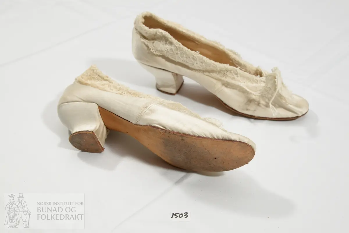 Sko trukket med hvit silkesateng. Påsydd kjøpeblonde i bomull langs åpning, sløyfe av blonde og silke på overlæret. Såle av skinn. Skinn i selve skoen. Snellehæl.