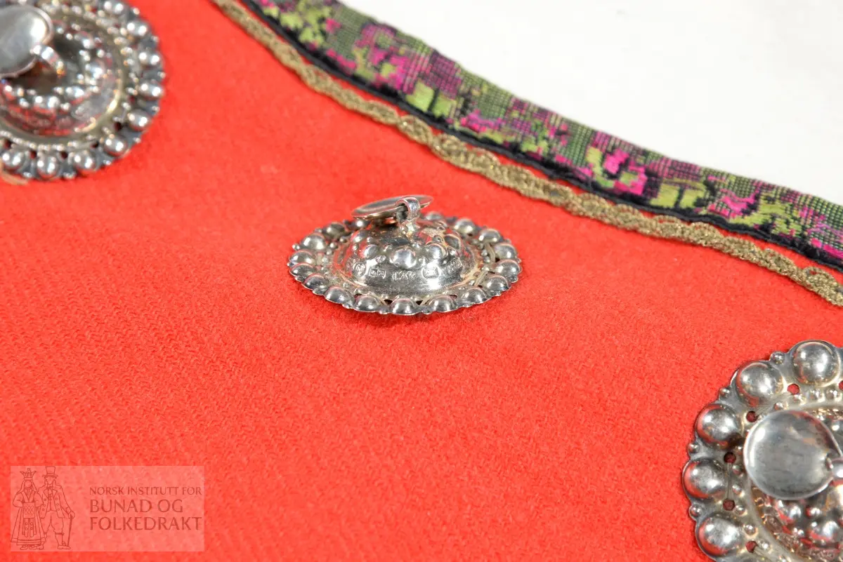 Liv sauma av raud ullkypert, lett valka. Fora med ubleika bomullslerret.
Ermhole, rundt hals og opning framme er kanta med blomstermønstra fleirfarga fløyelsband med svært avslitt fløyel. Innanfor kantingar er det påsett eit smalt metallband.
Etter framkantane er det påsett 3 borer på kvar side, dei to øverste litt breiare enn dei fire nederste, alle har eit skåleforma lauv festa i midten. På framsida av den kurva delen er det fem ulike stempel: 63 i eit sirkelforma stempel, truleg årstalsstempel. Månadsstempel: 6 over M i rektangelforma stempel. Mesterstempel i bølgeforma ramme LAR, Lorentz Christopher Angel Reimers, fekk borgarskap i Bergen i 1832. Lødighetsstempel 13 1/3. Trapesforma stempel med 7 prikkar, Bergens bystempel.
Lukking framme med to metallhekter på kvar side.
Heile livet er sauma på maskin. Kanting er sauma for hand. 
Livet sauma på stakken for hand. 
Stakken er av heimevove ullerret, blåsvart. Splittopning midt framme. Samansett av 5 rette høgder med jare i begge sider, alle høgder. Faldelagt oppe med eit glatt felt framme mot splitt. Høgdene er sauma saman med maskin. 
Nedkant: 3 svarte smale sillkefløyelsband, nedanfor dei eit påsett stoffstykke av svart bomullsfløyel. Dette stoffet er ein hengefald, ikkje sauma fast i nedkant.
Nedkanten er skonna på innsida med eit lerretsvove mohairstoff av mørk grøn farge. Sauma på med maskin. I nedkant av skonninga er det påsett eit blåsvart kosteband sauma på for hand.
Kanten på linninga er av svart bomullssateng. Er bretta dobbelt og sauma på for hand på begge sider