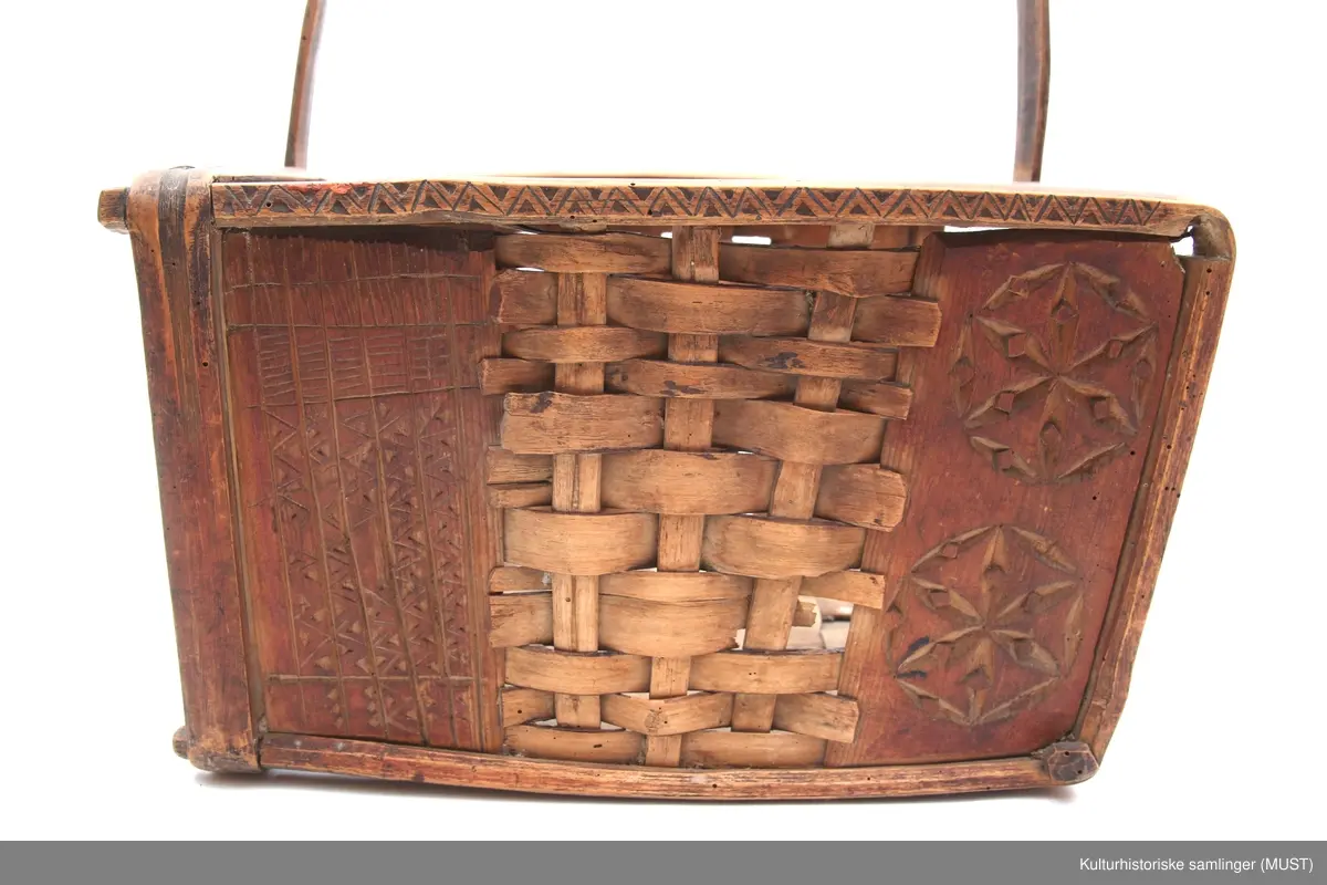 Firkantet trekurv med flettedekor og treskjæring på både kurv og håndtak. Dekoren består av to seksbladete blomster på hver side samt ulike trekantmønstre langs kanten av kurven og håndtaket. En av sidene i kurven mangler.