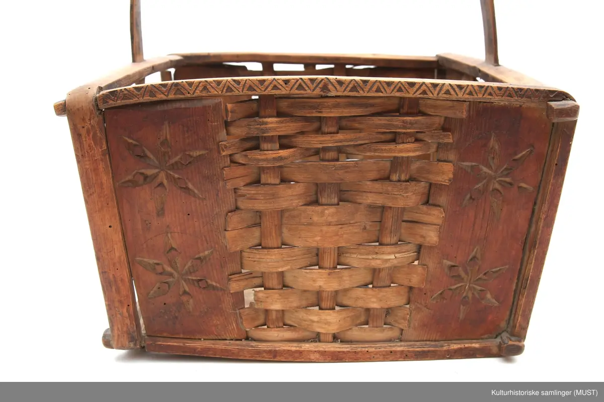 Firkantet trekurv med flettedekor og treskjæring på både kurv og håndtak. Dekoren består av to seksbladete blomster på hver side samt ulike trekantmønstre langs kanten av kurven og håndtaket. En av sidene i kurven mangler.