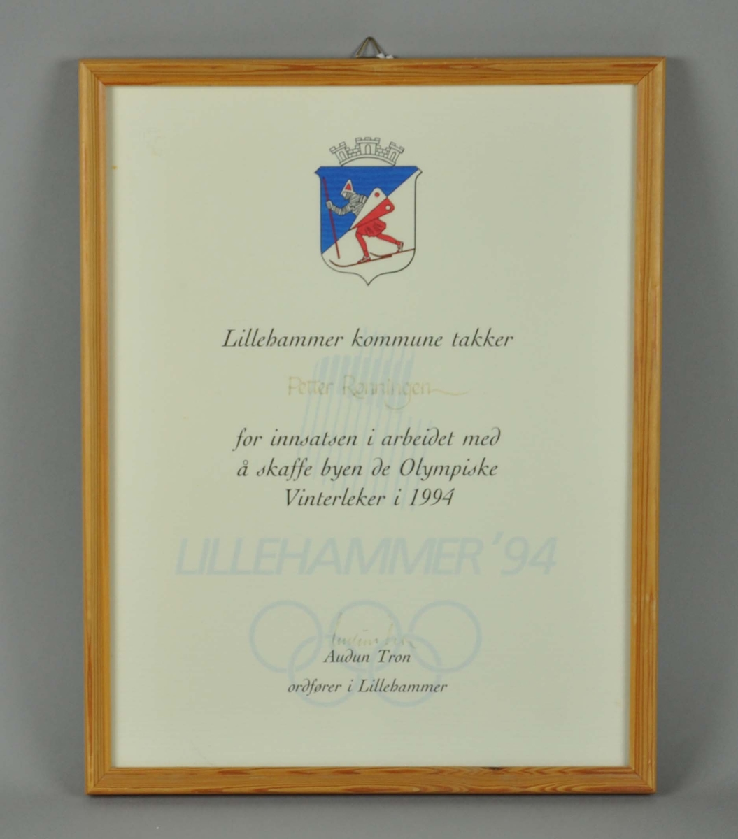 Innrammet diplom med Lillehammer kommunes byvåpen øverst og tekst med Lillehammer'94 logo og de olympiske ringer som bakgrunn under teksten.