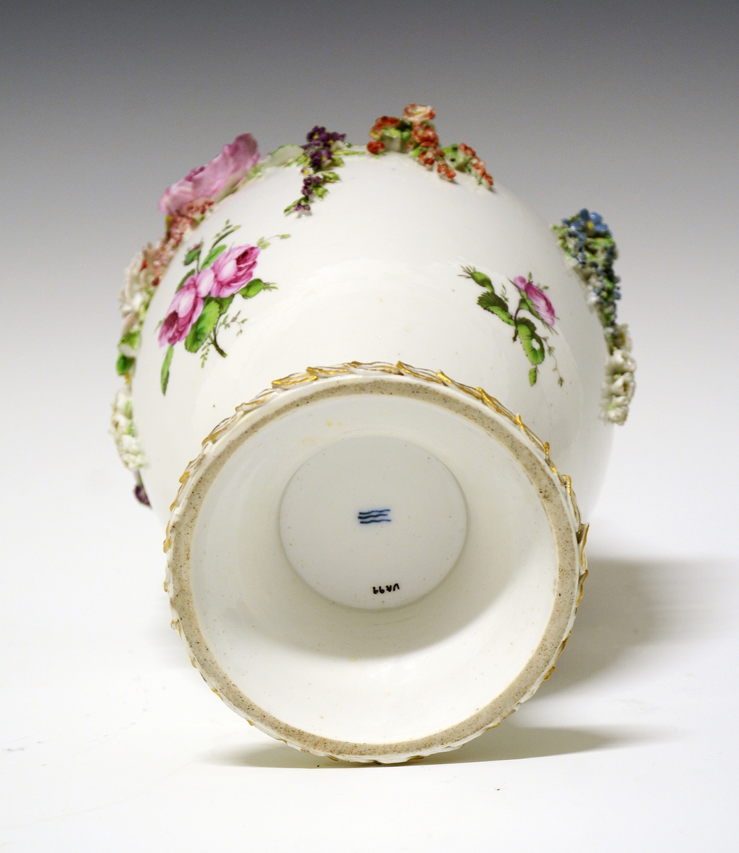 Potpourrikrukke av porselen med stett og lokk. Lokket er dekorert med håndmalt blomsterdekor og langs kanten en bord med blader i tre høyder dekorert med gull, over bladene gullfargede små kuler. På toppen av lokket en kjerub eller kupid som sitter på sitt ene kne. Han har på seg et rosa lendeklede og har holdt i et skjold eller en atributt. 
Urnens korpus er dekorert med håndmalte blomster, og modellerte blomster som sitter i klynger og strekker seg nedover korpus. De modellerte blomstene er uglasert og håndmalt. Urnen mangler en hanke. Den eksisterende hanken har modellerte blomster ved festene som strekker seg nedover i klynger.  Langs stettens kant samme bladbord som på lokket.