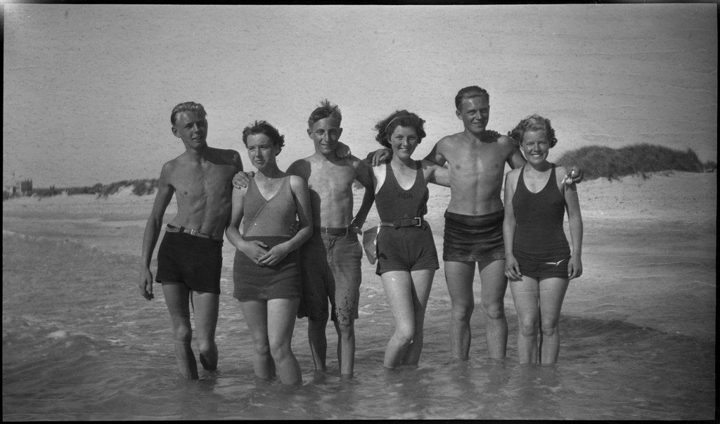 En stor vennegjeng i lystig lag på stranda på Sola. De har trommer og trekkspill og leker i vannet og på land med og uten ball. Sola strandhotell ligger i bakgrunnen. Tor Johannessen og Harald Bergsaker er på bilde nr. 14 og 18.