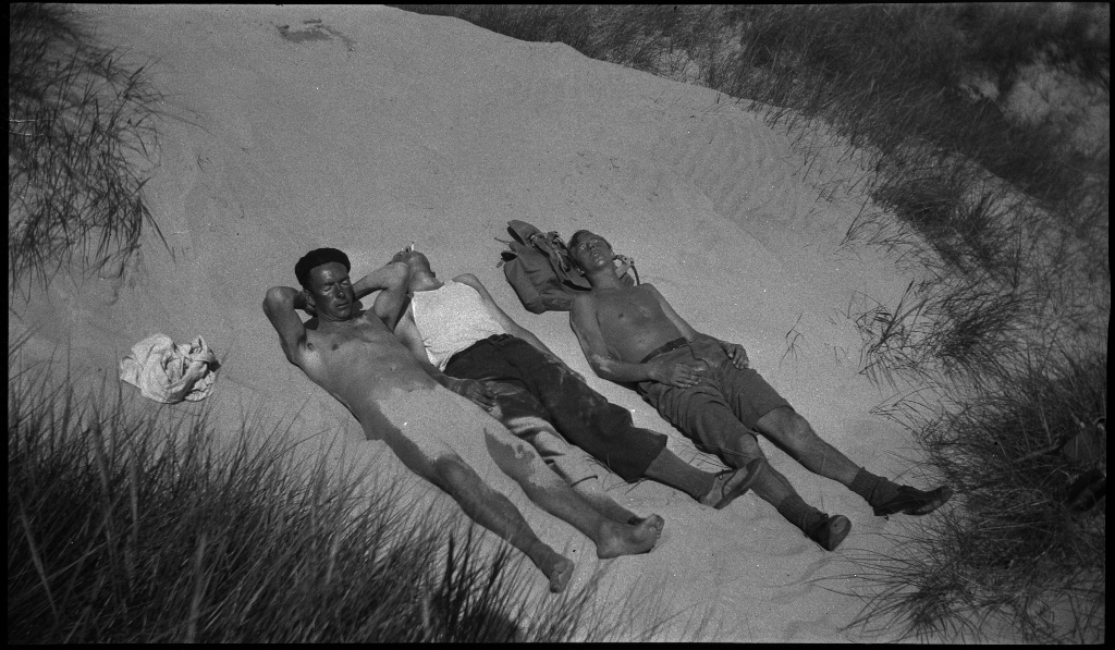 Unge menn og gutter soler seg i sanddynene på Solasanden. To gutter leker med en kikkert. Lindtner har svart alpelue på.